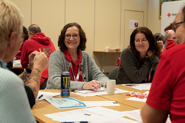 Flere personer sitter rundt et bord. To damer sitter med ansiktet mot kamera og ansiktene deres er i fokus, de smiler til de andre som sitter med ryggen til rundt bordet