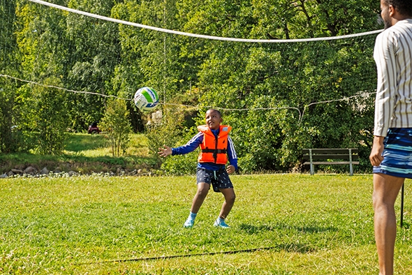 Bildet er av en volleyballbane. En gutt strekker seg etter en volleyball. Du ser ryggen til pappaen og litt av nettet i tillegg til gutten.
