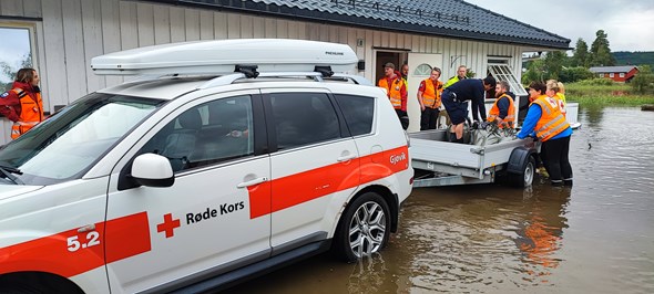 Bilde av en bil som står i vann utenfor et hus. Mange frivillige fra Røde Kors henter ting ut fra det oversvømte huset