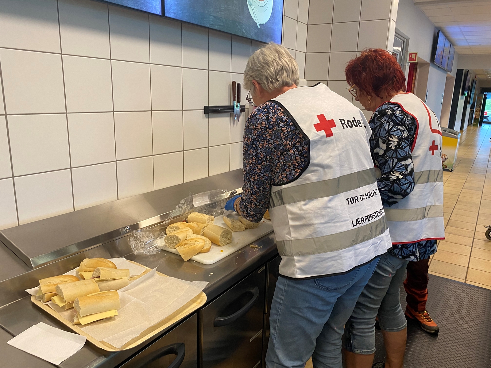 To damer står med ryggen til på et storkjøkken. Det ligger baguetter på disken som smøres. Damene har Røde kors-vester på seg.
