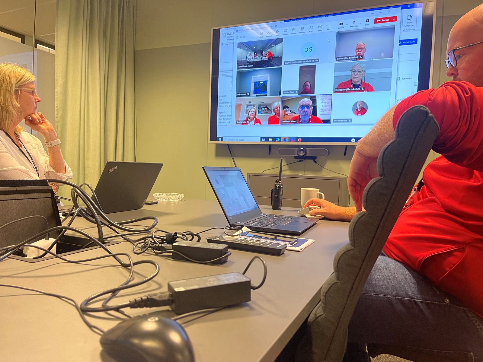 Bildet viser en mann og en dame som sitter i teammøte med flere personer på en stor skjerm mellom dem.