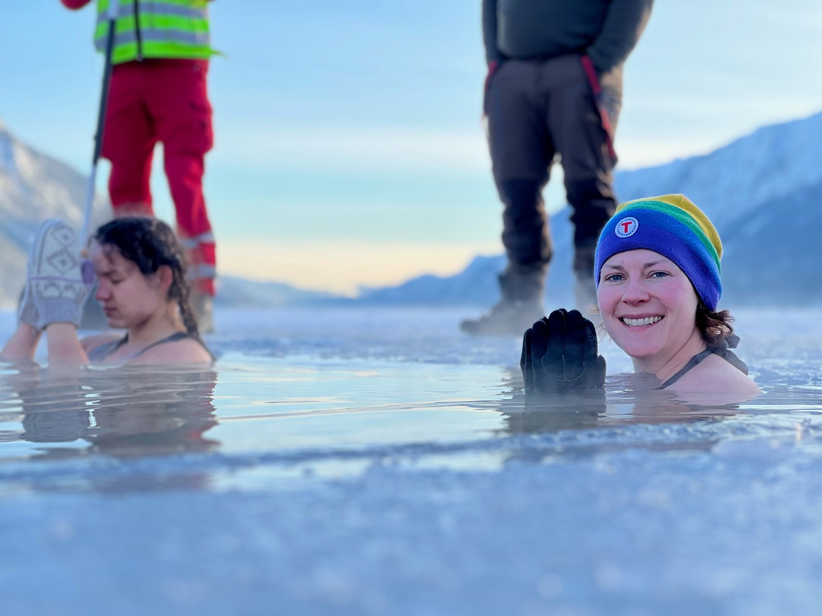 To damer sitter i vannet på en isdekt innsjø. Ser to bein i bakgrunnen som står og passer på. 