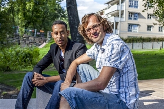 Bildet viser to menn som sitter på en benk. De smiler mot kamera. Sola skinner.