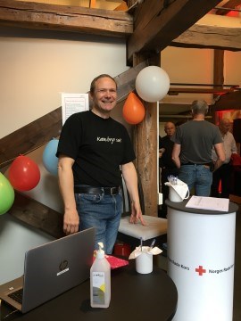 Bildet viser en av Fellesverkets frivillige, Georg. En voksen mann smiler til kamera. Det henger ballonger på veggen bak ham.