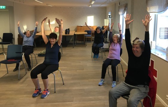 Bildet viser et knippe eldre damer som trener sittende på hver sin stol. De strekker armene i været og smiler.
