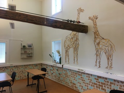 Bildet viser et klasserom med pulter og stoler. På veggene er det malt dekorative tegninger, blant annet av to store sjiraffer.