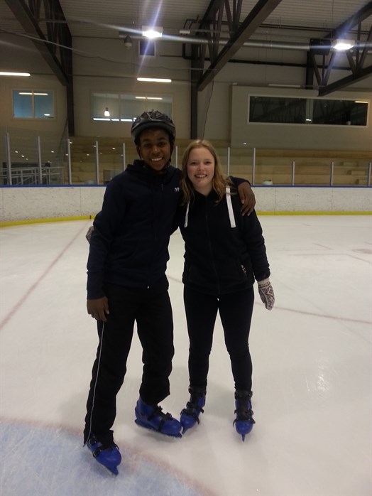 Bildet viser to ungdommer med isskøyter på. De står på isen i skøytehallen og smiler til kamera.