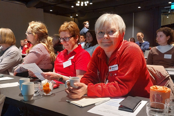 Bildet viser en kvinne sitte i en forsamling. De lytter til foredragsholdere. Hun er kledt i rødt.