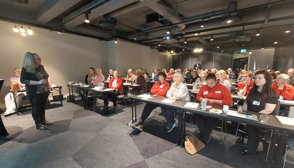 Bildet viser en stor forsamling på 69 personer som lytter til foredragsholdere på scenen. Mange er kledt i rødt.