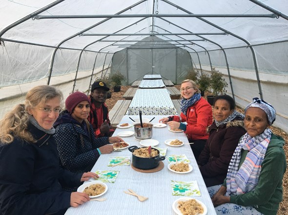 Bildet viser frivillige, deltakere og ansatte rundt et bord. Bordet er plassert i et drivhus. De nyter et deilig varmt måltid sammen.