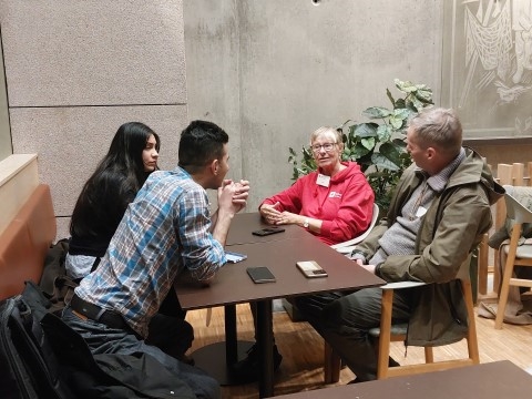 En gruppe mennesker sitter sammen rundt et bord og prater. De smiler og har det hyggelig.