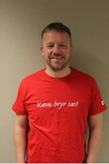 Odd-Christian Lilleeng ser mot kameraet og smiler, i en rød tskjorte med påskriften "Kæm bryr sæ?". Foto.