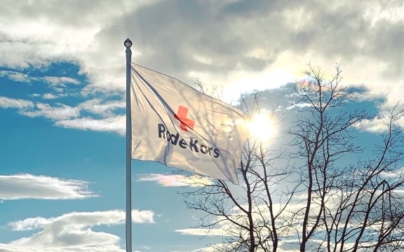 Bildet viser Røde Kors-flagget som vaier i vinden. Det er sol og noen få skyer på himmelen.