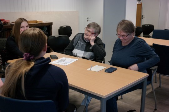 Bildet viser to pensjonister sammen med to ungdomsskoleelever. Elevene viser de eldre hvordan de kan bruke nettbrettet og smarttelefonen sin.
