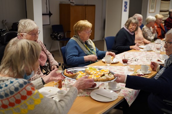 Bildet viser et gjeng eldre som sitter sammen rundt et bord. De spiser snitter og drikker kaffe sammen.