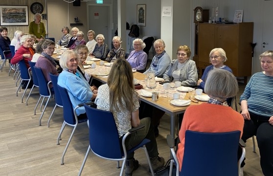 Bildet viser en stor forsamling på circa 25 personer som sitter rundt et bord. Det er dekket på med kaffekopper og fat.
