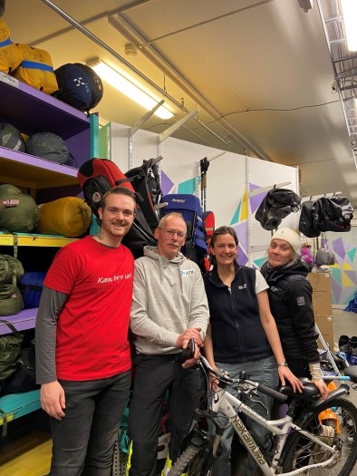 På bildet ser vi fire personer smiler til kamera. De har på arbeidsklær. De står foran hyller med ulikt turutstyr.