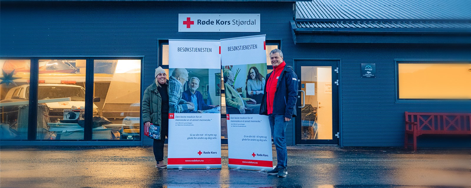 Ansatt og aktivitetsleder for besøkstjenesten i Stjørdal poserer med roll-ups utenfor Røde Kors-huset.