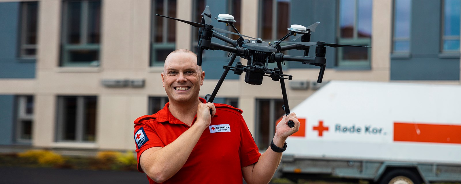 Mann i hjelpekorps-uniform poserer smilende. Holder drone på skulderen.