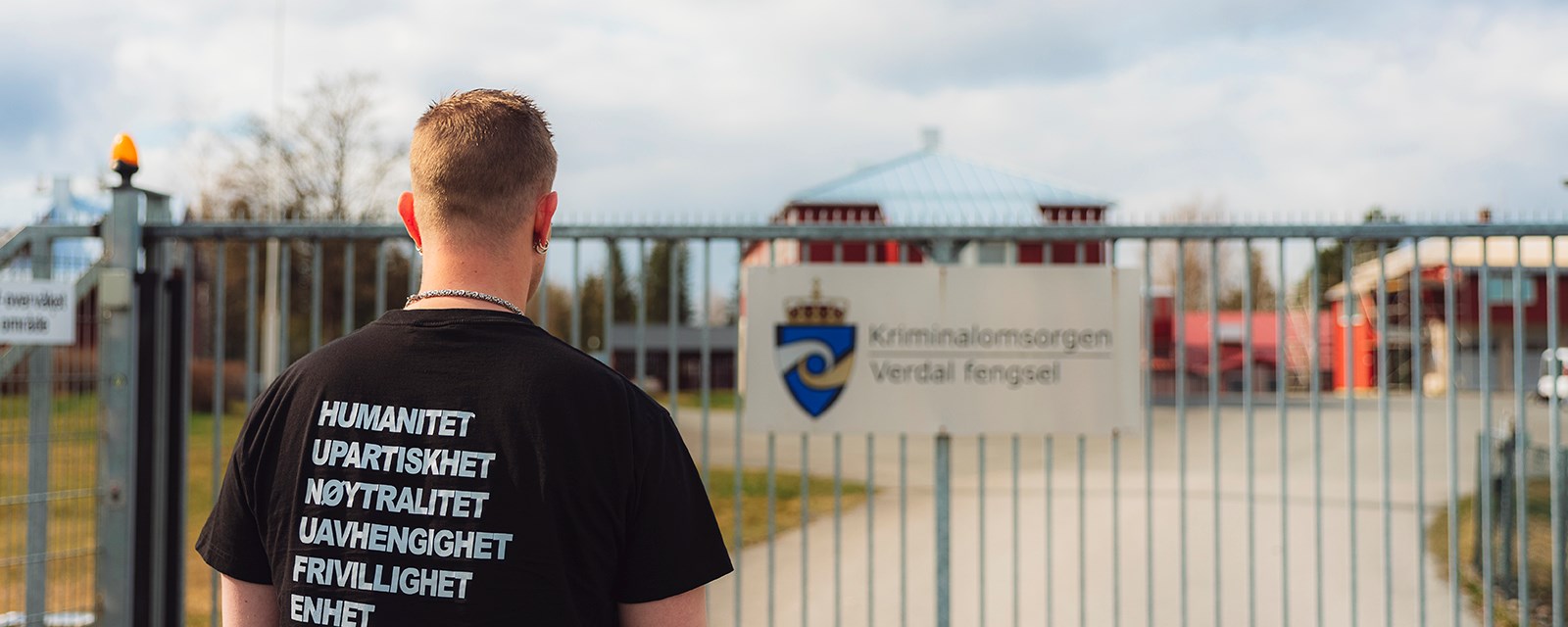 Innsatt med ryggen til foran porten til Verdal fengsel. Svart t-skjorte med Røde Kors-prinsippene trykket på ryggen.