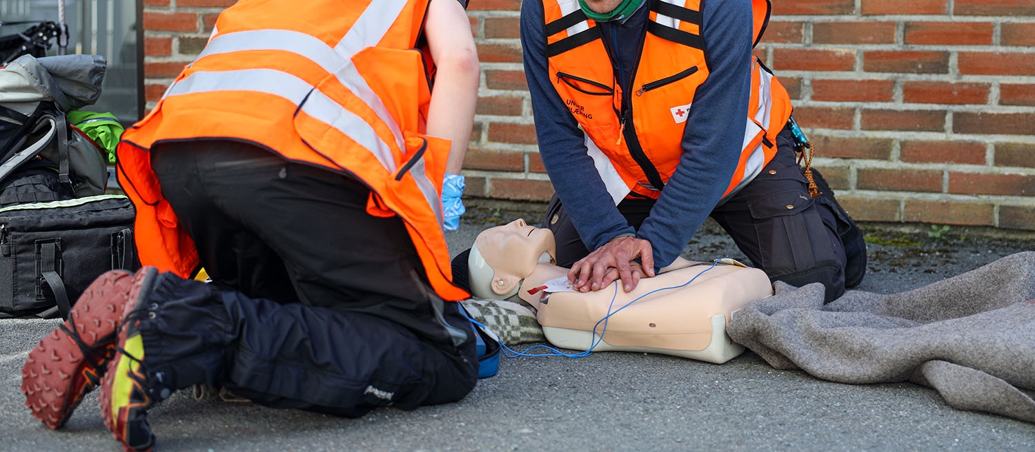 To menn i hjelpekorpsvester utfører hjerte- og lungeredning på en dukke som ligger på bakken