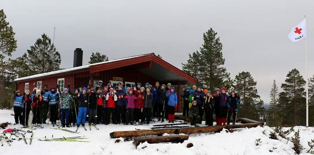 Bilde av hytte, stor folkemengde foran og et flaggstang med Røde Kors-flagg, i snødekt landskap