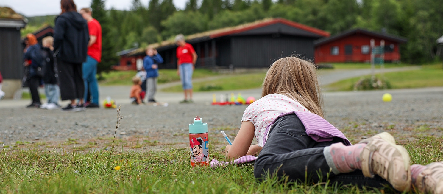 En ung jente ligger på gresset hvor hun tegner, samtidig som vi ser lekende foreldre og barn i bakgrunnen