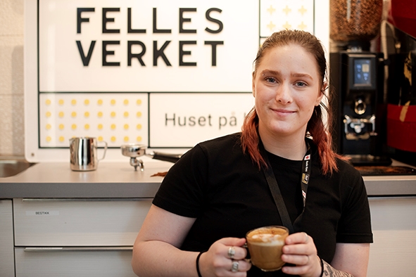 En ungdom står med en selvlaget barista-kaffe foran Fellesverket-logoen