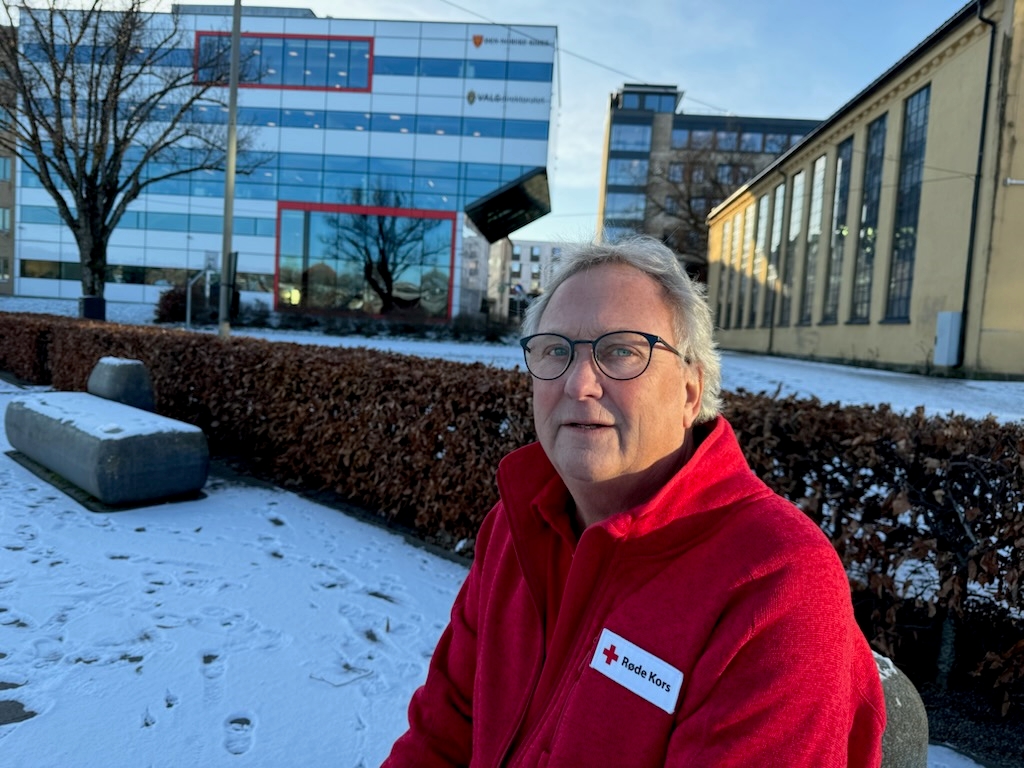 Portrett av en mann i Røde Kors-klær som sitter ute. Det er bygninger i bakgrunnen og snø på bakken.