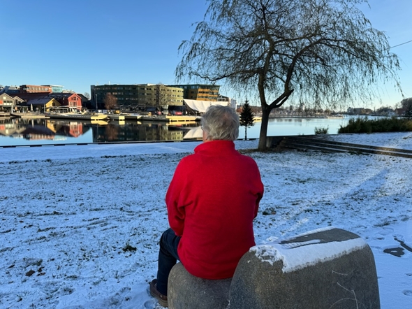 En mann sitter på en stein med ryggen til og titter ut på elva. Det er snø og blå himmel.