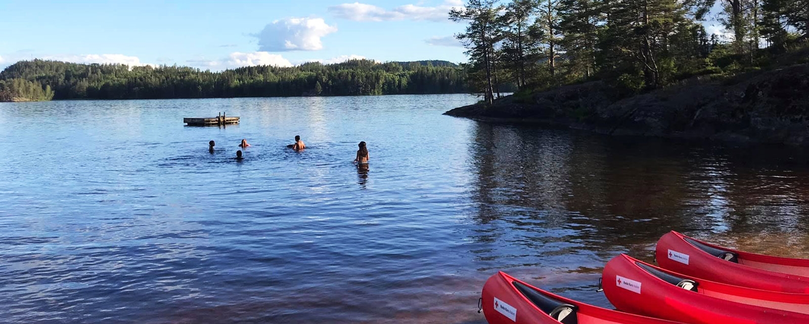 Åpent vann med folk som bader.  I hjørnet av bilde på land så er de tre røde kanoer. Det er en varm skyfri sommer vær.