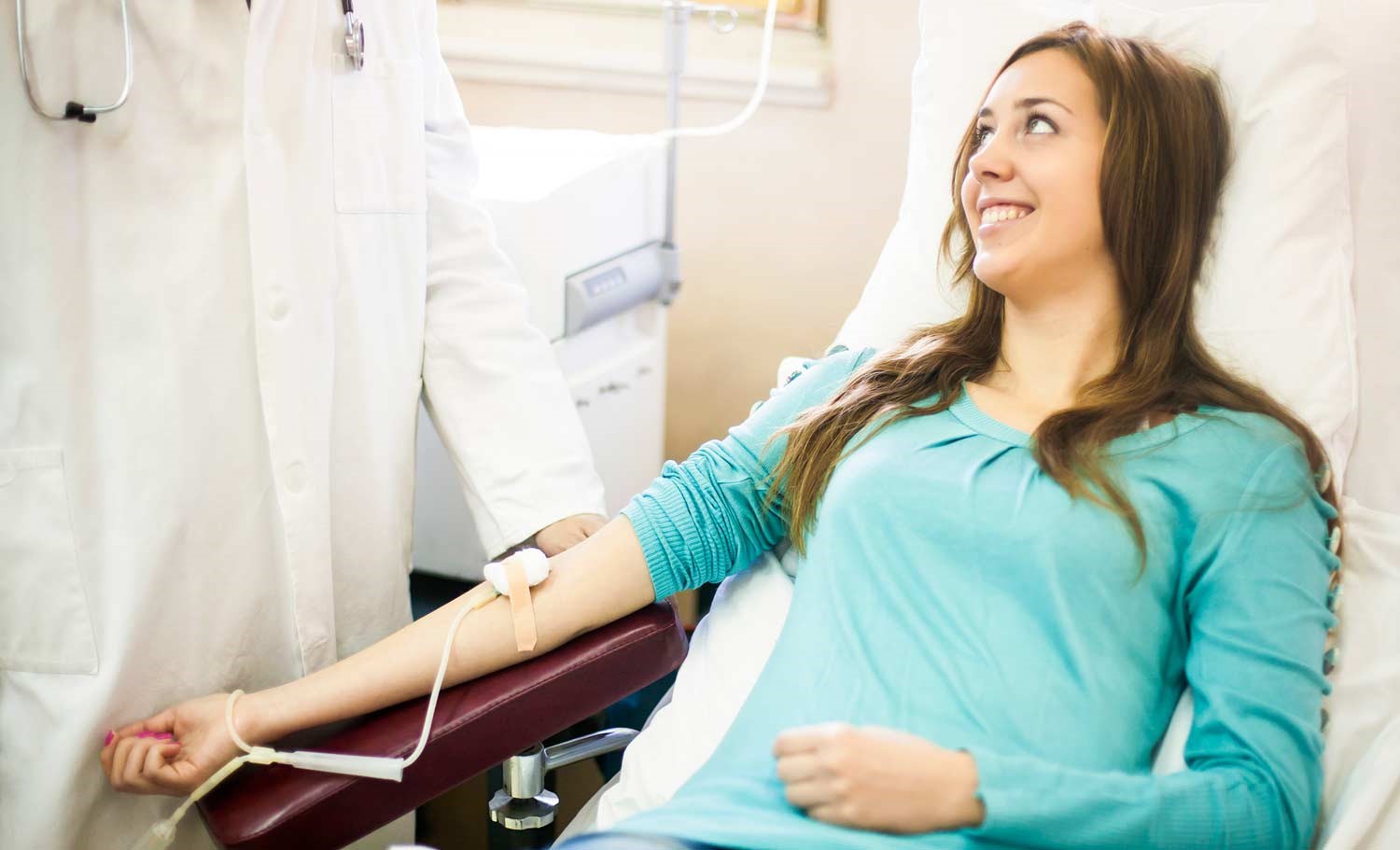 En ung kvinne med langt mørkt hår sitter smilende i en blodgiverstol mens hun tapper blod