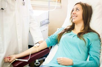 En ung kvinne med langt mørkt hår sitter smilende i en blodgiverstol mens hun tapper blod