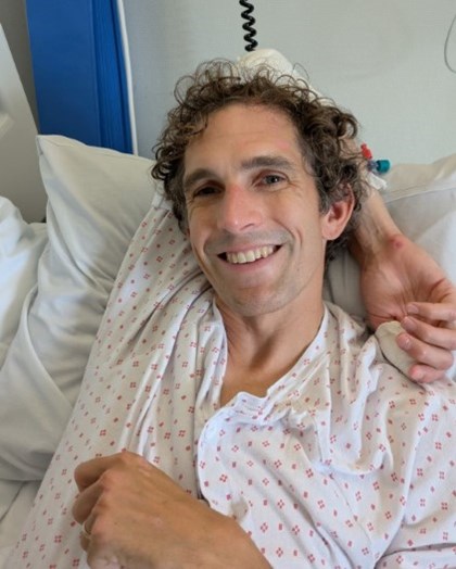 En mann ligger skadet i en sykeseng men smiler og er ved godt mot.