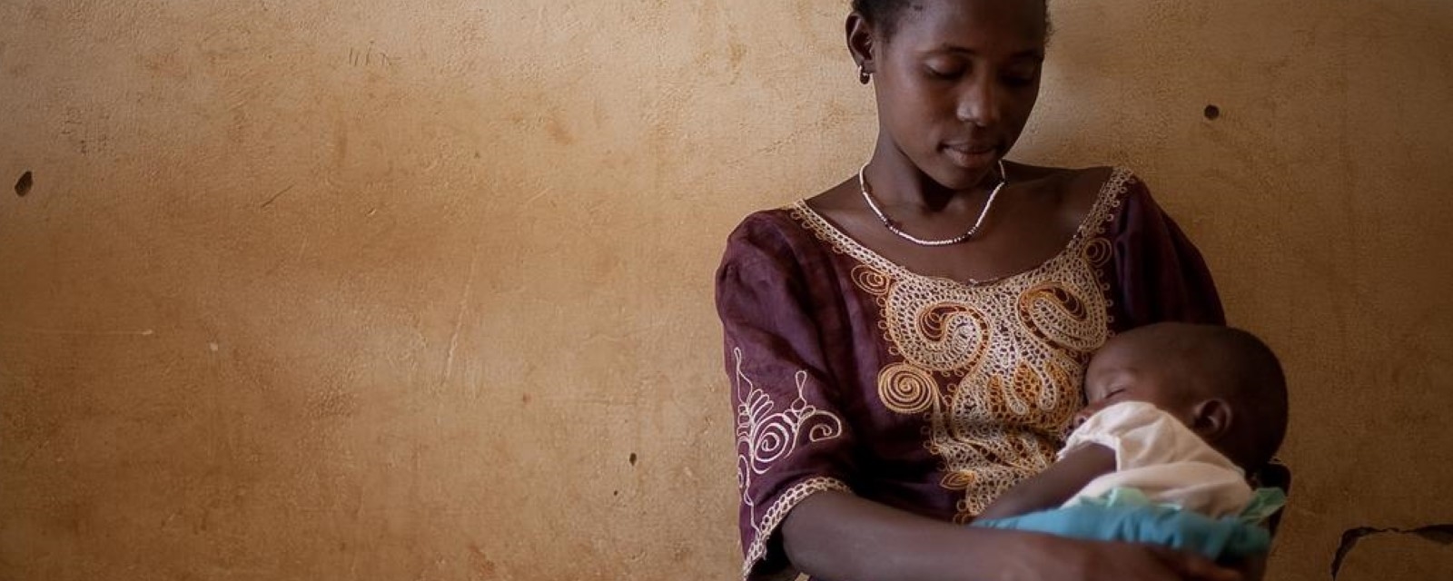 En kvinne sitter mot en vegg og ser ned på babyen hun holder i armene sine.