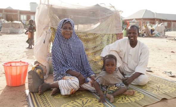 En familie på tre sitter på et teppe utenfor et lite telt i en flyktningleir.