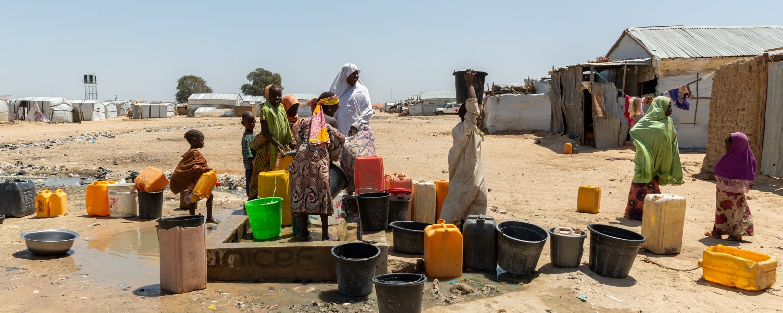 Voksne og barn i en flyktningleir er samlet rundt en vannkran og fyller på bøtter og kar.