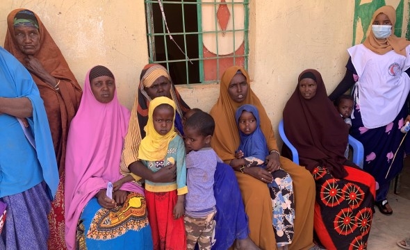 Flere kvinner og barn sitter utenfor et murhus og venter på helsesjekk.