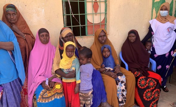 Flere kvinner og barn sitter utenfor et murhus og venter på helsesjekk.