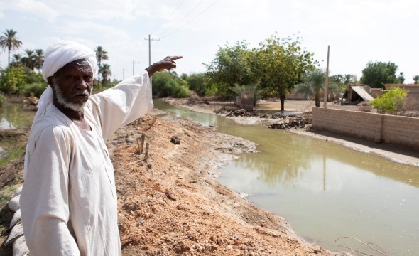 En mann med hvit kjortel peker mot et hus som ligger i et området rammet av flom.