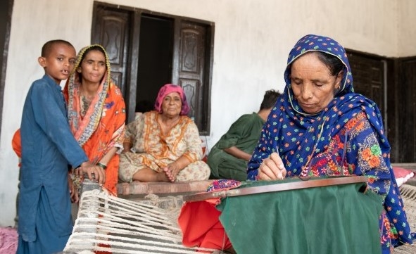 En kvinne sitter i en seng og broderer mens tre andre ser på.