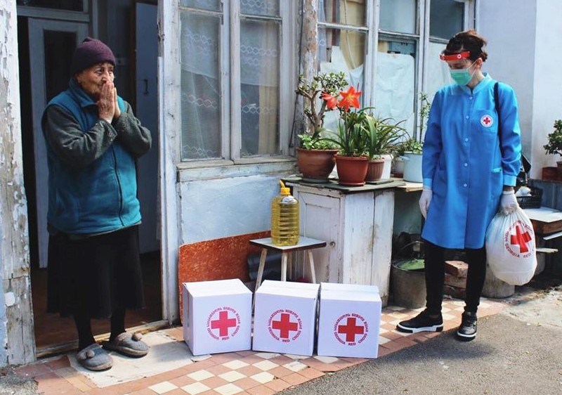 En eldre kvinne står utenfor huset sitt. Ved siden av henne står en frivillig i smittevernsutstyr med tre store hvite bokser med Røde Kors-logo på.