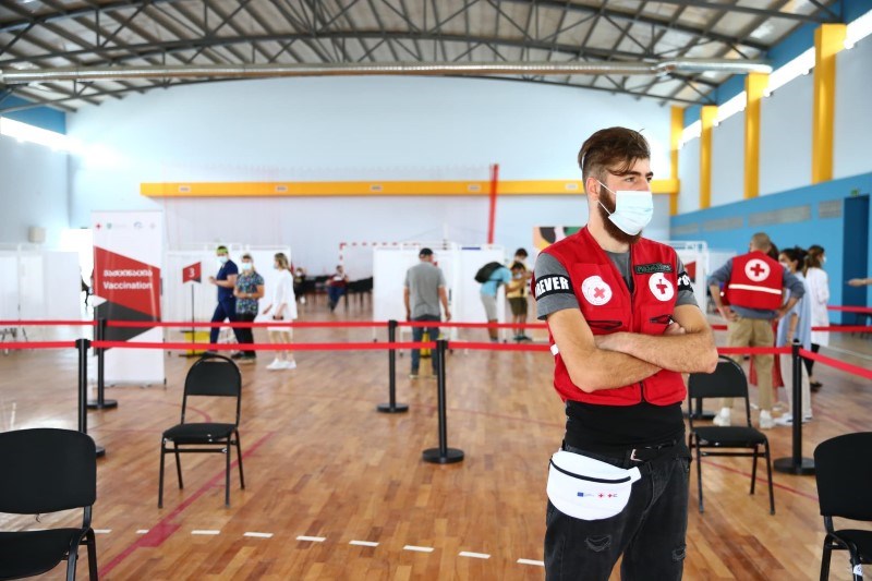 En mann med munnbind og Røde Kors-unfiform står i en idrettshall som er omgjort til vaksinesenter.