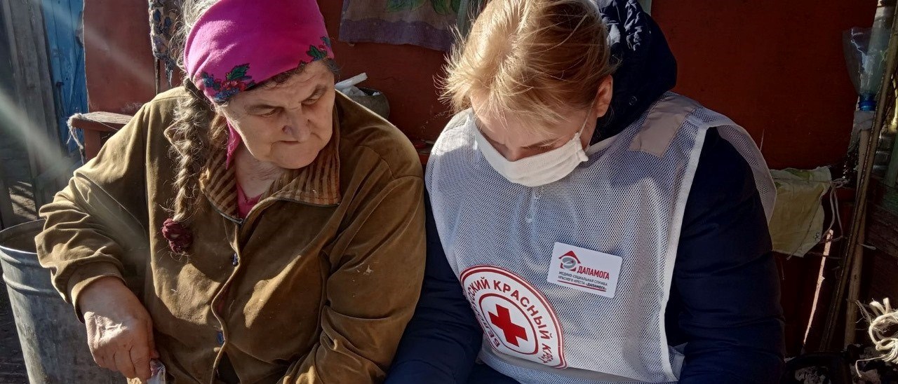 En eldre kvinne sitter ved siden av en kvinnelig frivillig fra Røde Kors som har på seg munnbind.