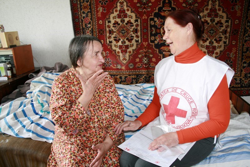 En eldre kvinne sitter på en seng med en kvinnelig frivillig fra Røde Kors. De snakker sammen og smiler.