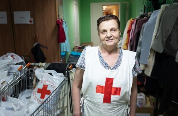 en eldre dame jobber som frivillig i Russland Røde Kors. Hun står i et  rom fullt av klær som hun deler ut til flyktninger.