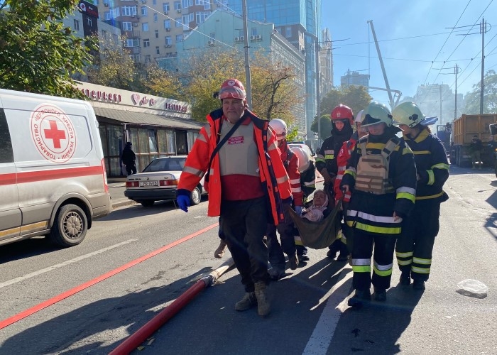 Seks personer bærer en skadet kvinne. Røyken stiger i bakgrunnen.