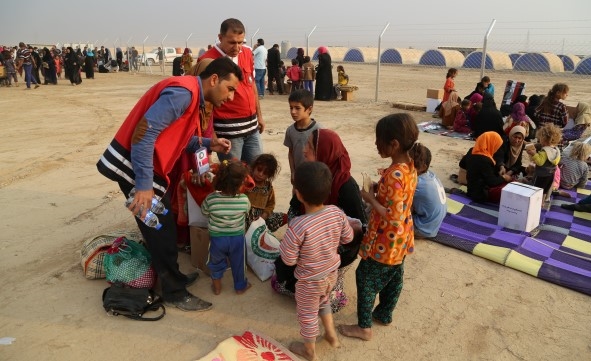 To frivillige deler ut vann og snakker med en kvinne i en flyktningleir. Rundt henne står det 5 små barn.