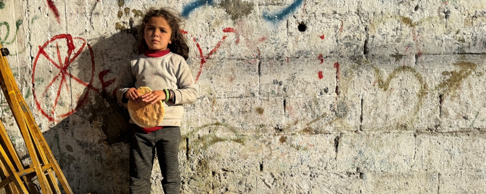 En liten jente står foran en murvegg med et brød i hånden.