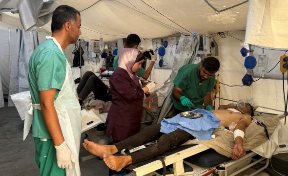 To mannlige sykepleiere undersøker en skadet pasient som ligger på en sykeseng med armene utslått. Ved siden av står en kvinnelig pårørende.